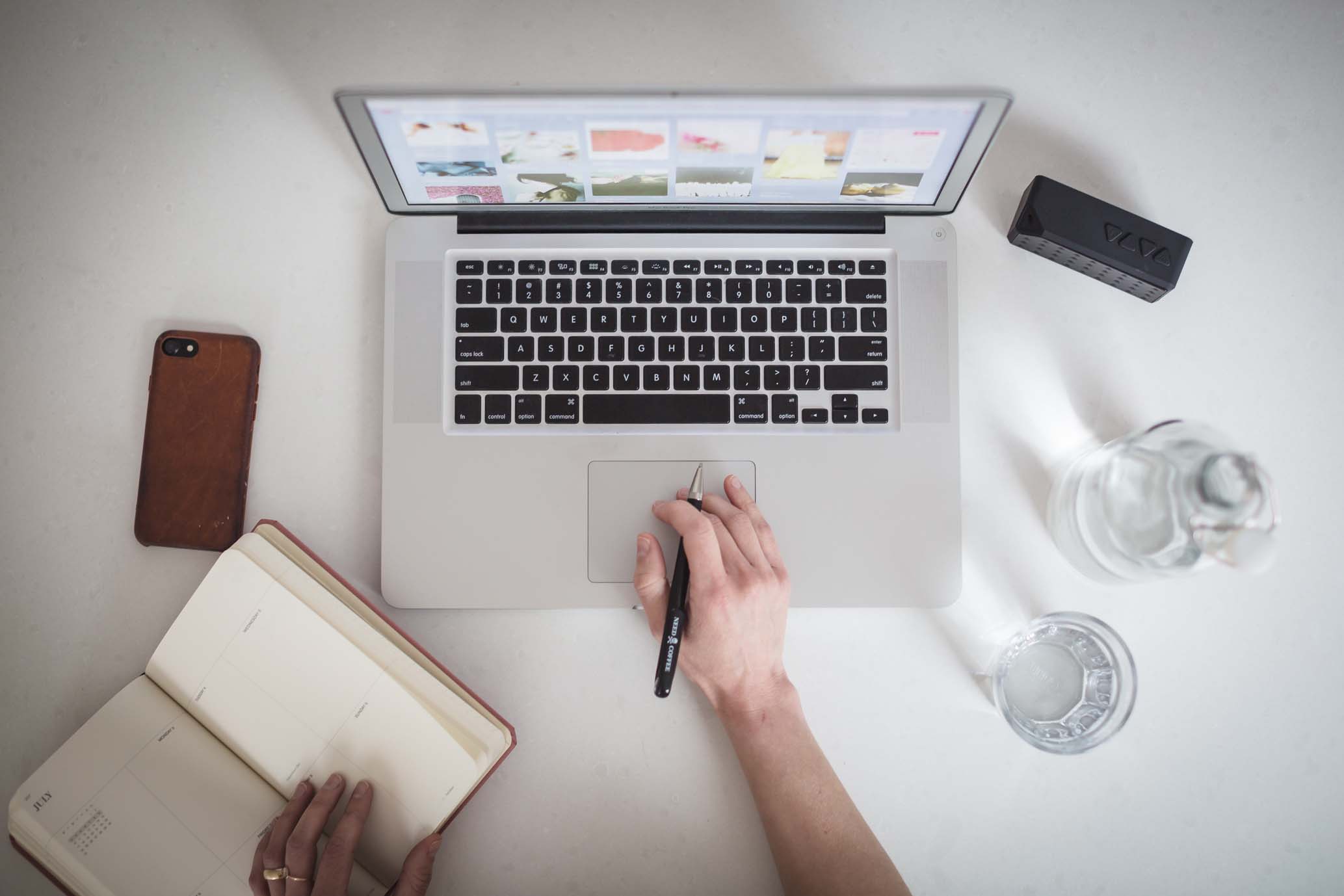 Overhead view of a laptop with a colorful screen, a smartphone, an open notebook, a hand holding a pen and a water bottle on a wooden desk, bathed in warm golden-hour light with soft amber and cream tones.